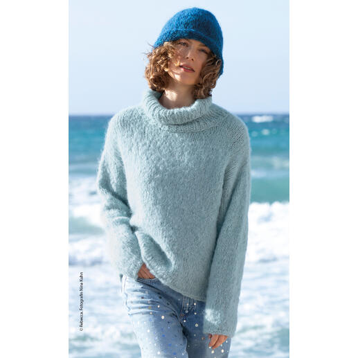 Frau in einem hellblauen Pullover mit hohem Kragen und Mütze, steht am Strand.