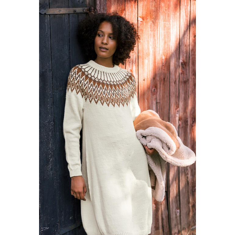 Frau in einem langen, hellen Kleid mit gestricktem Muster, hält einen Pullover in der Hand.