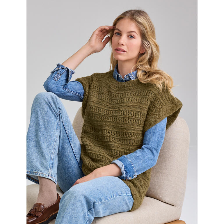 Frau sitzt auf einem Stuhl, trägt einen ärmellosen Pullunder und eine Jeans, schaut zur Seite.