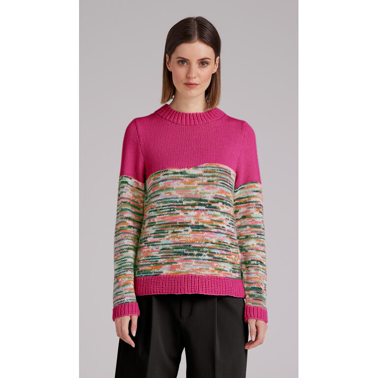Frau trägt einen bunten Pullover mit geometrischem Muster, steht vor einem neutralen Hintergrund.