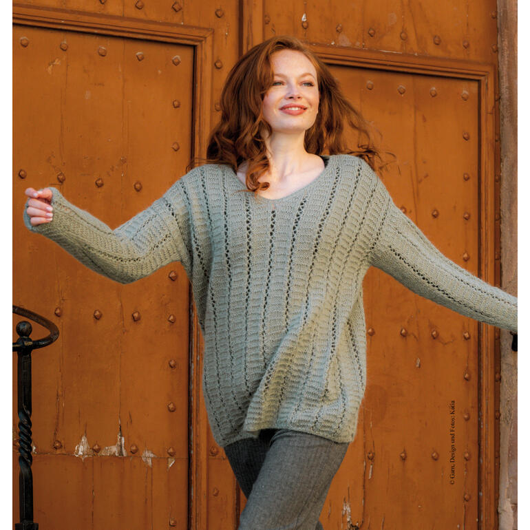 Frau in einem hellgrauen Pullover aus Soft Gratté, vor einer rustikalen Holztür.