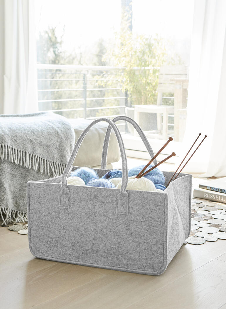 Helle Filztasche mit Wolle und Stricknadeln im Innenraum, auf einem Holzboden.