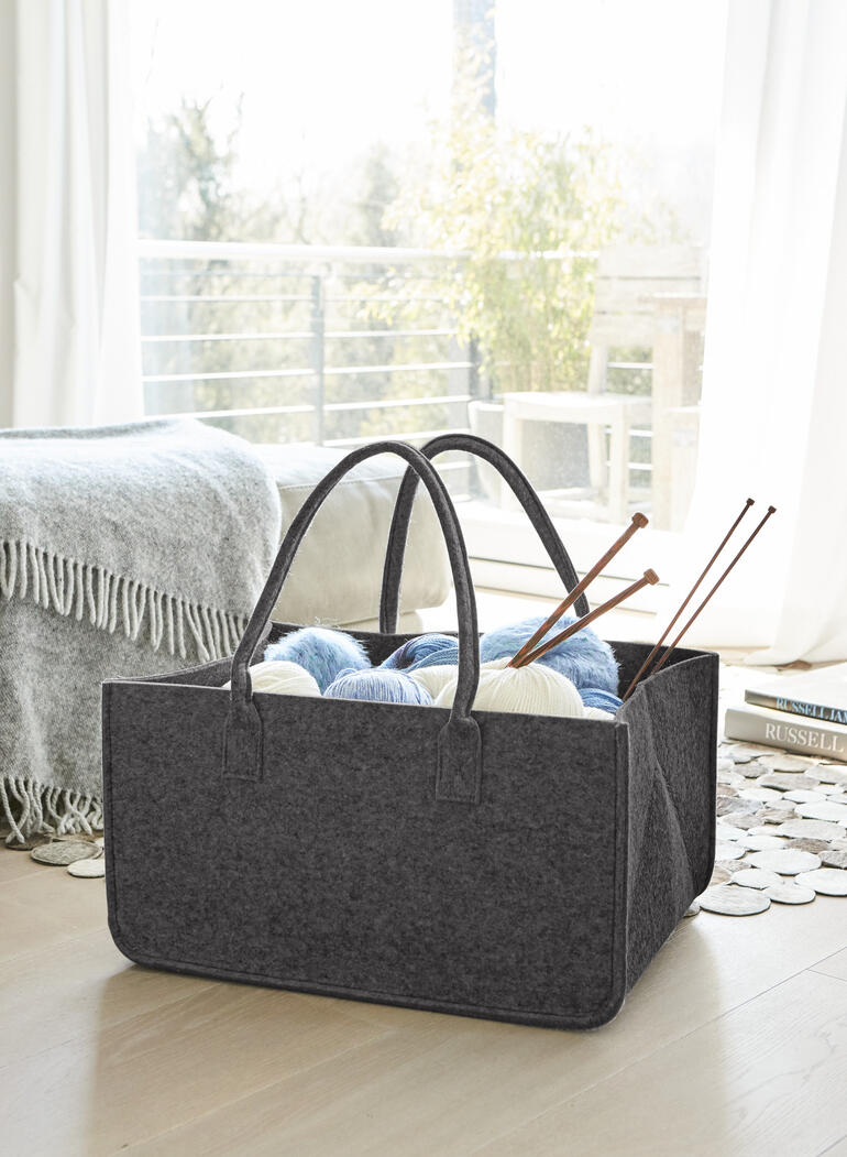Dunkelgraue Filztasche mit Wolle und Stricknadeln im Innenraum, auf einem Holzboden.