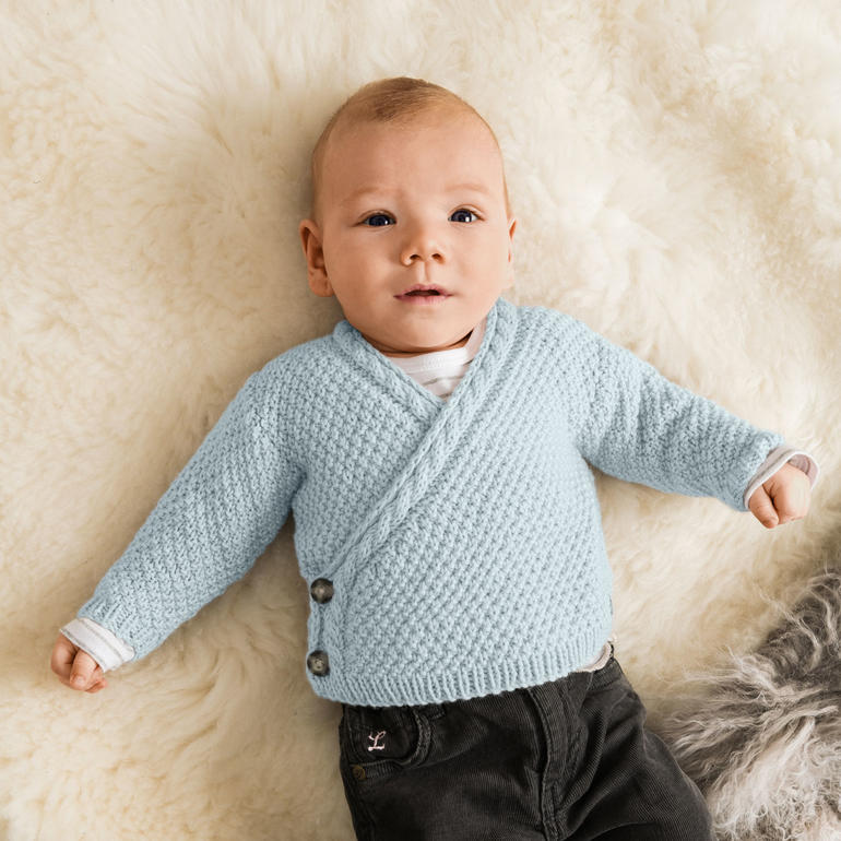 Baby in blauer Wickeljacke mit Knöpfen auf einem weichen Untergrund.