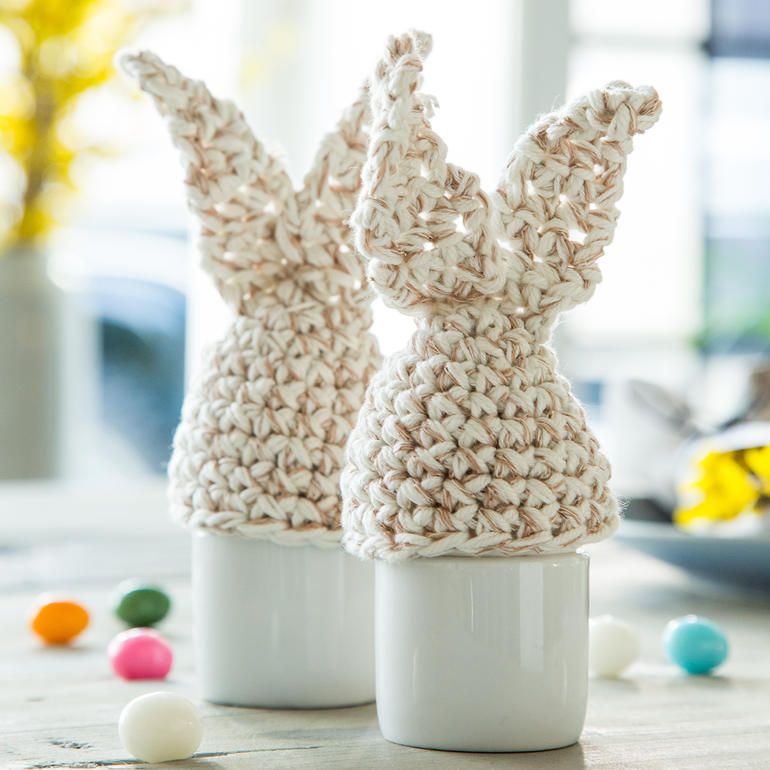 Zwei Eierwärmer in Form von Hasenohren aus hellem Garn, auf einem Tisch.