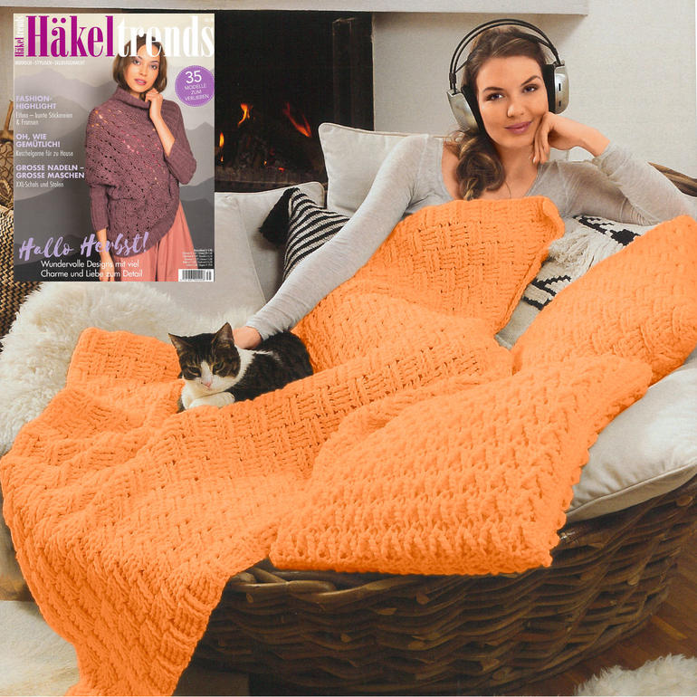 Frau sitzt auf einem Sofa, eingehüllt in eine orangefarbene Häkeldecke, mit Katze auf ihrem Schoß.