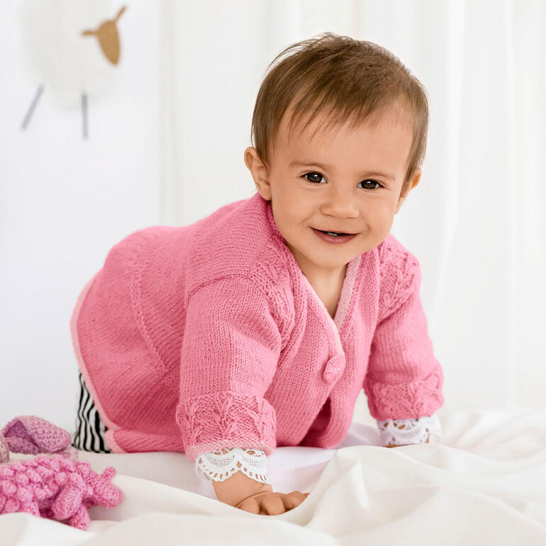 Baby krabbelt in einer rosa Babyjacke aus Baby Merino, lächelt freundlich.
