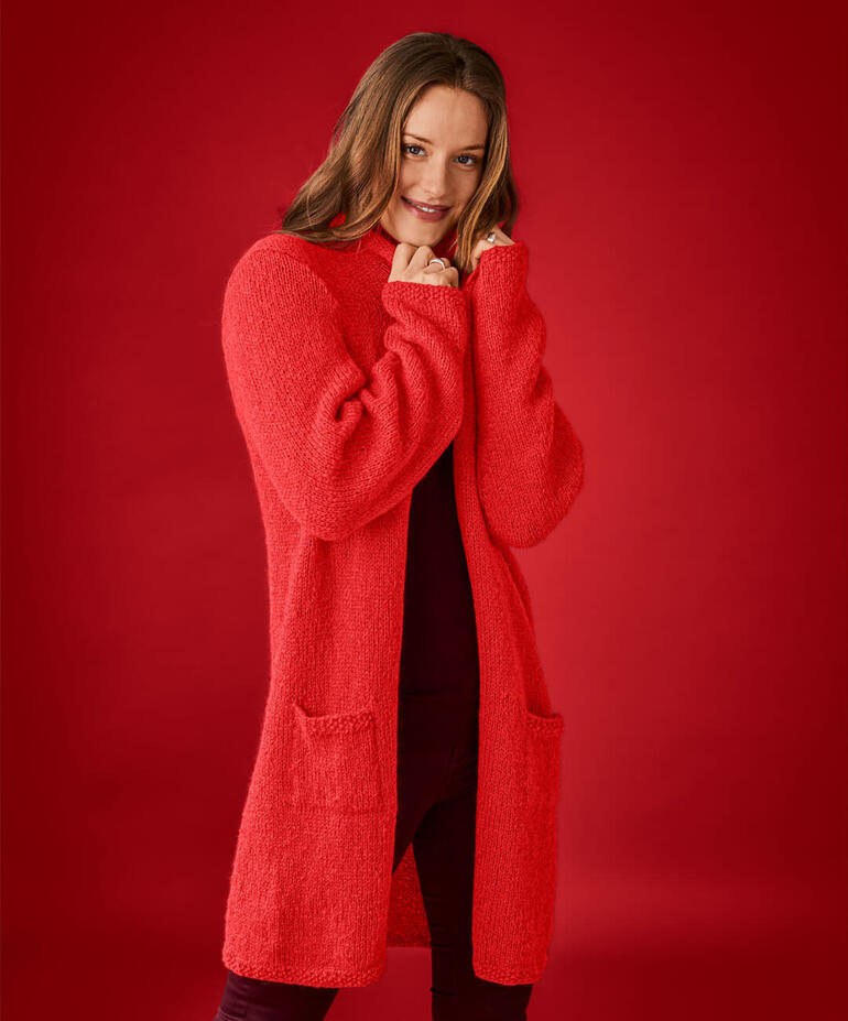 Frau in roter Longjacke aus Alpaka Deluxe posiert vor einem roten Hintergrund.