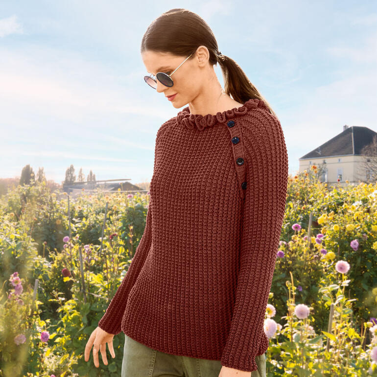 Frau in einem roten Pulli mit Knöpfen auf der Schulter, umgeben von Blumen.