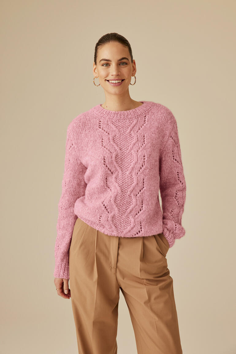 Frau in rosa Strickpullover und beiger Hose posiert lächelnd in einem hellen Raum.