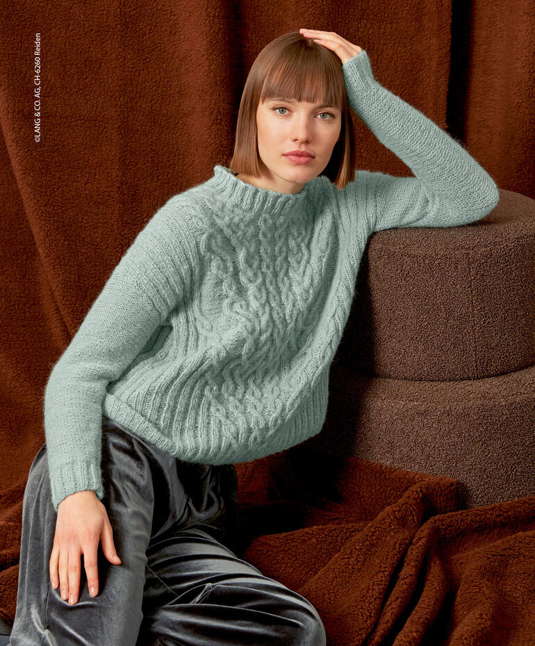 Frau in mintgrünem Pullover lehnt lässig an Kissen auf einem braunen Hintergrund.