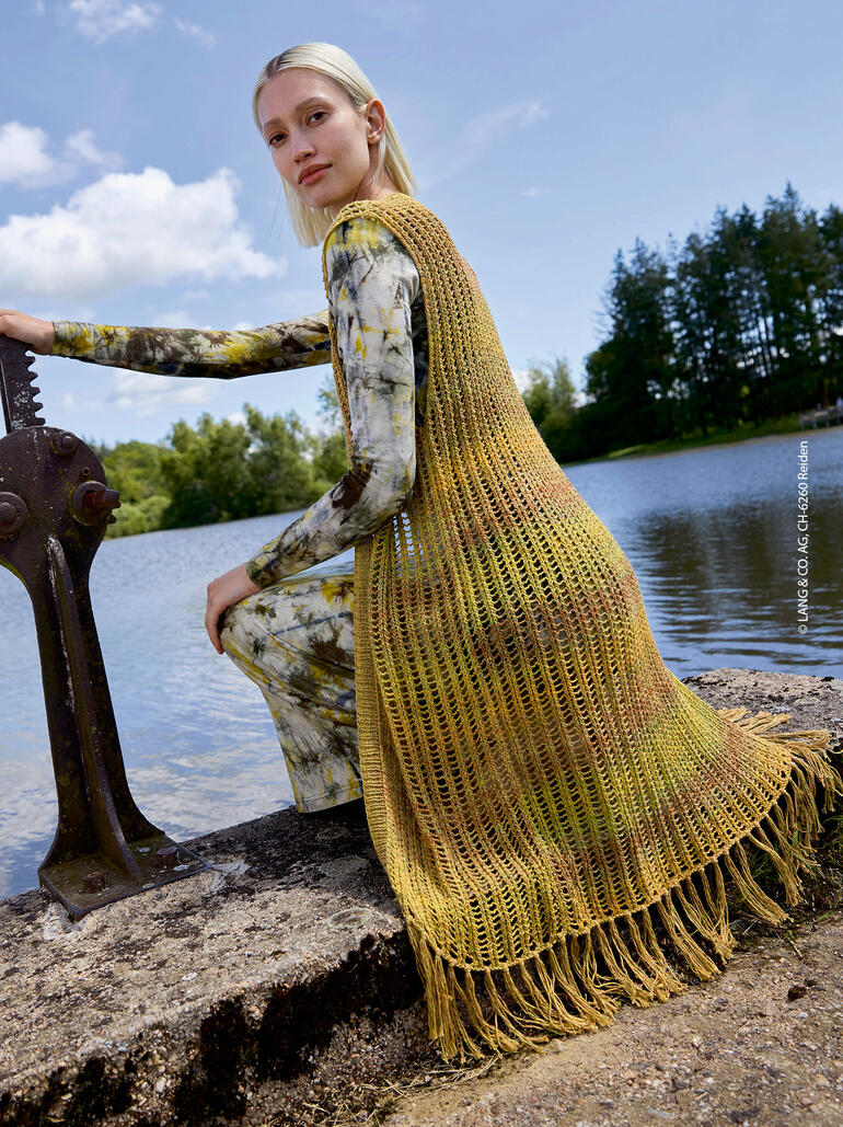 Frau in einer langen, offenen Weste sitzt am Ufer eines Sees.
