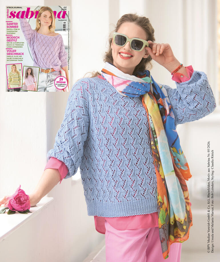 Frau in einer blauen, luftigen Strickjacke hält eine Blume und posiert.