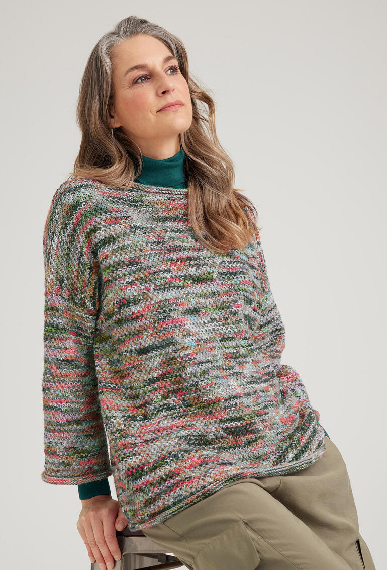 Frau sitzt in einem bunten Merino-Pullover auf einem Hocker.