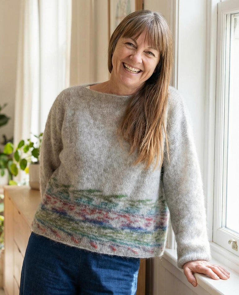 Frau lächelt in einem grauen Pullover mit buntem Muster, steht vor einem Fenster.
