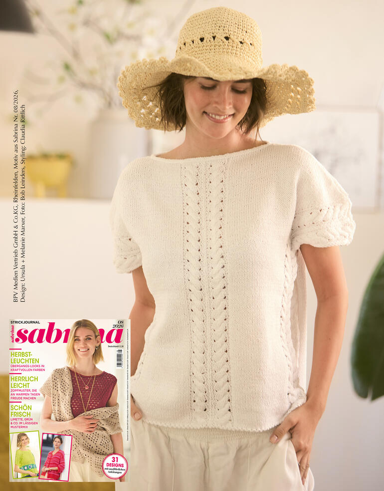 Frau trägt weißen Pulli aus Cottonprima, steht mit Strohhut im Hintergrund.
