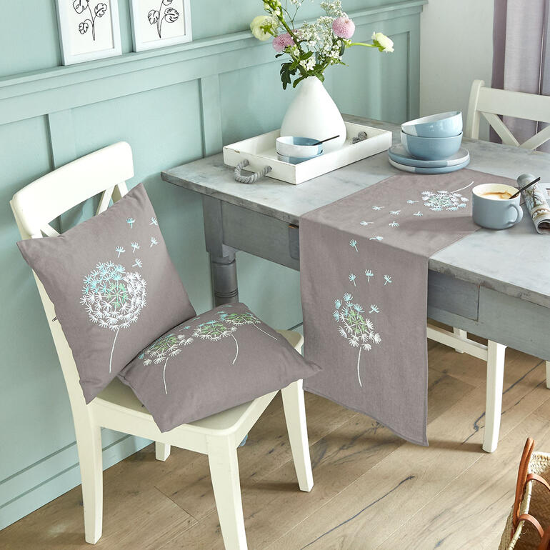 Tischläufer und Kissen mit Pusteblumenmotiven auf einem Tisch.